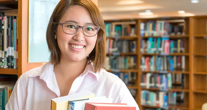 A young librarian in glasses holding books in a library.