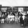 Children sitting on chairs in a library listening to a librarian tell a story